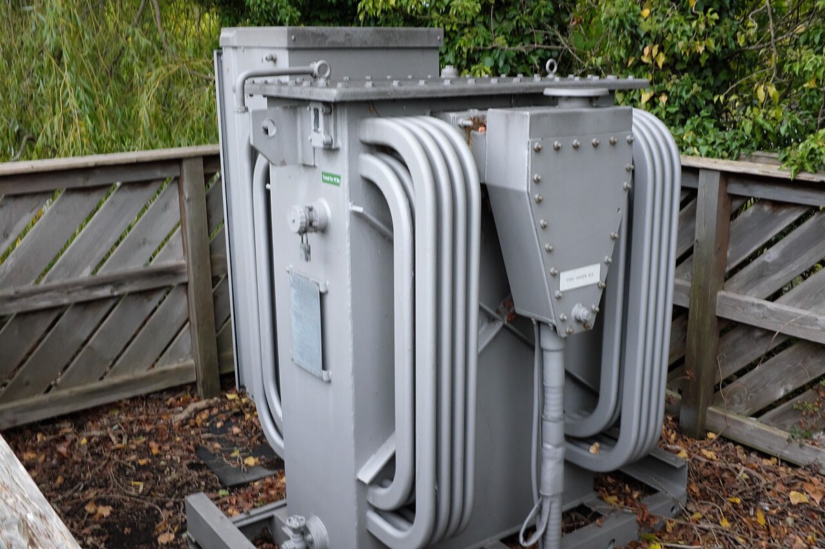 A grey metal transformer with curved cooling pipes inside a wooden fence