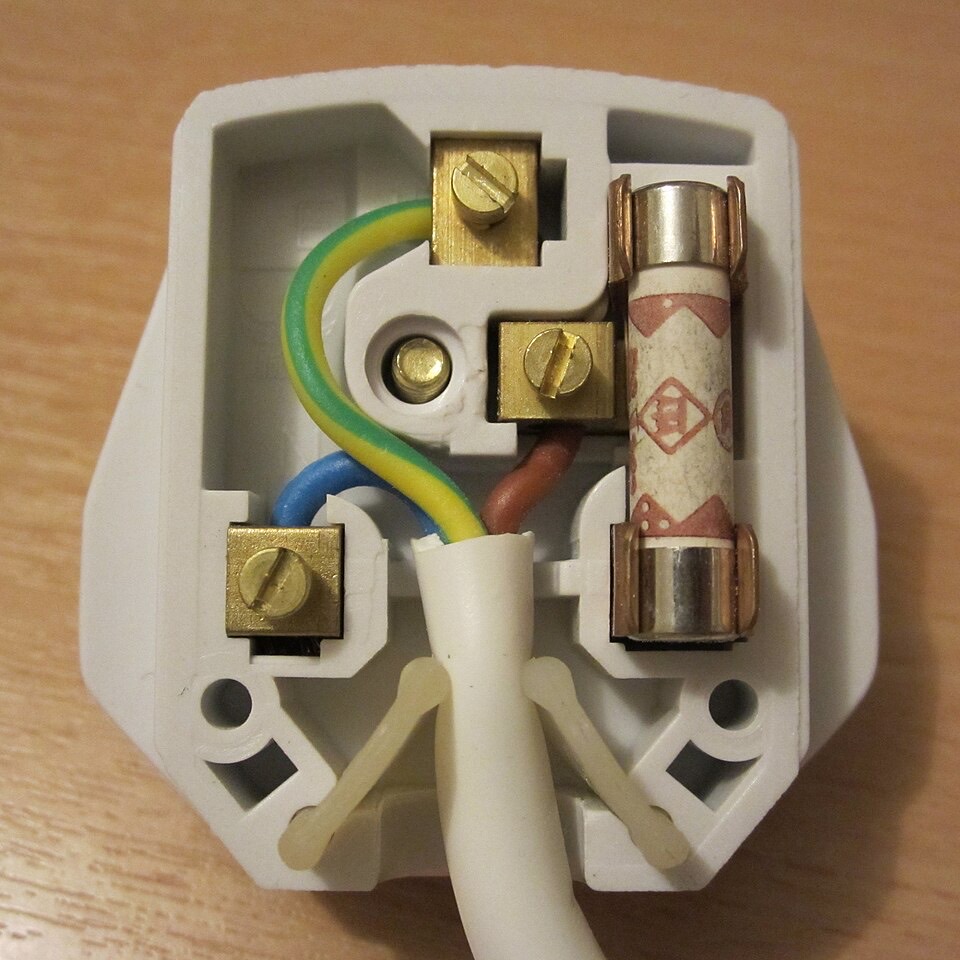 An open UK three-pin plug: a green and yellow wire runs to the top pin, a blue wire to the bottom left pin and a brown wire to a fuse on the right