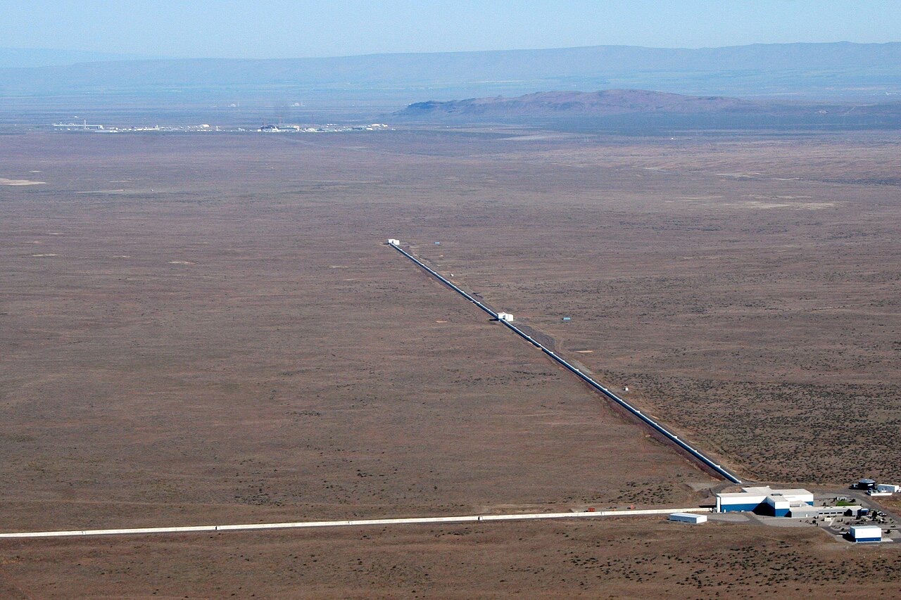 Aerial view of a flat desert with two long straight pipes meeting at a right angle at a group of buildings