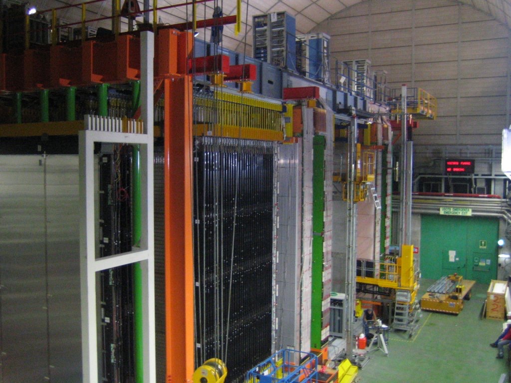 A hall-sized particle detector with tall racks of equipment, gantries and a yellow lift