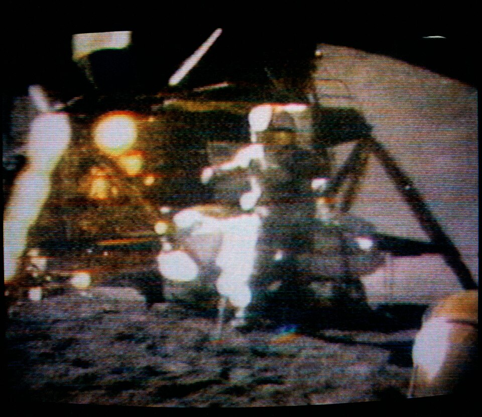 A blurred color television picture of an astronaut in a white spacesuit standing on the lunar surface in front of the lunar module