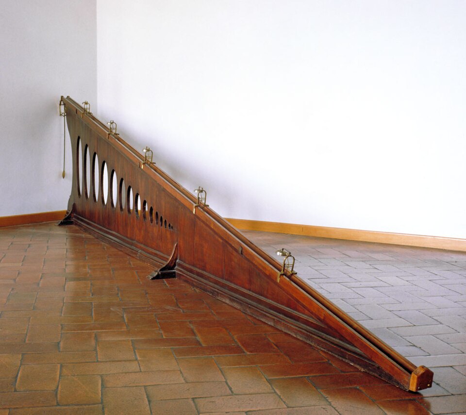 A long wooden ramp with a groove along its top edge and five small brass bells mounted along it at increasing distances