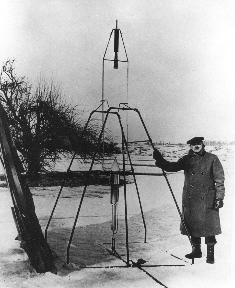 Black and white photograph of Robert Goddard in a long coat and cap, standing in a snowy field beside a tall metal frame that holds a slender rocket