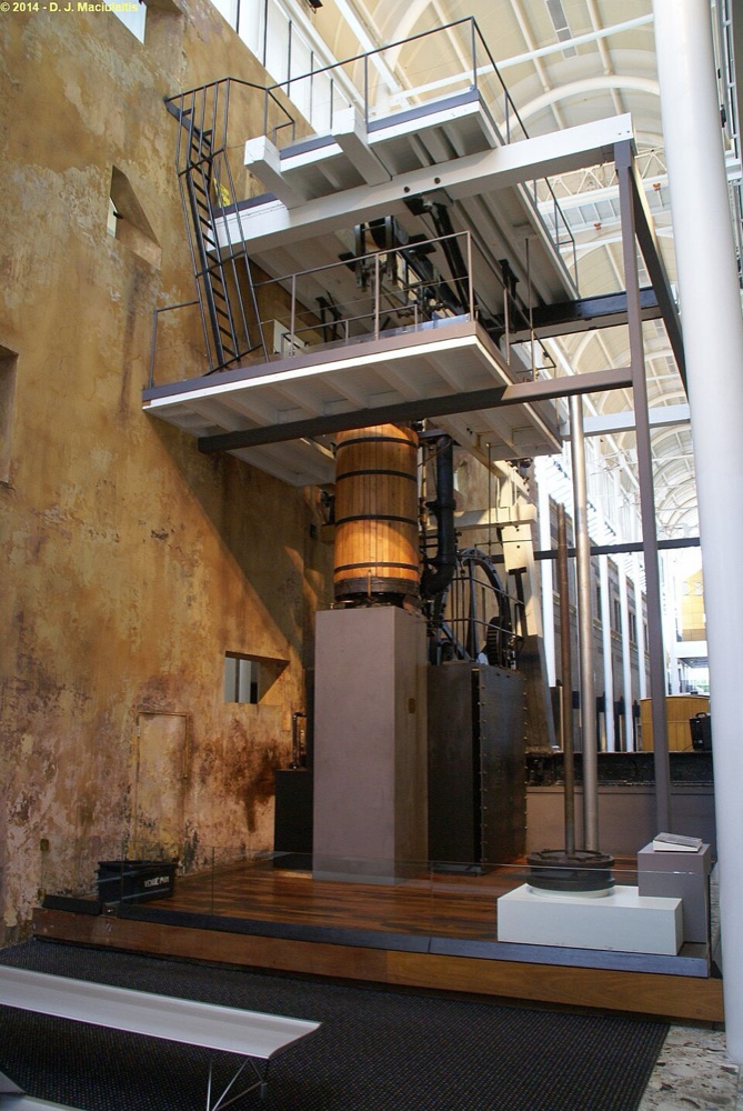 A large steam engine in a museum, with a wooden beam overhead, a tall vertical cylinder and a big spoked flywheel