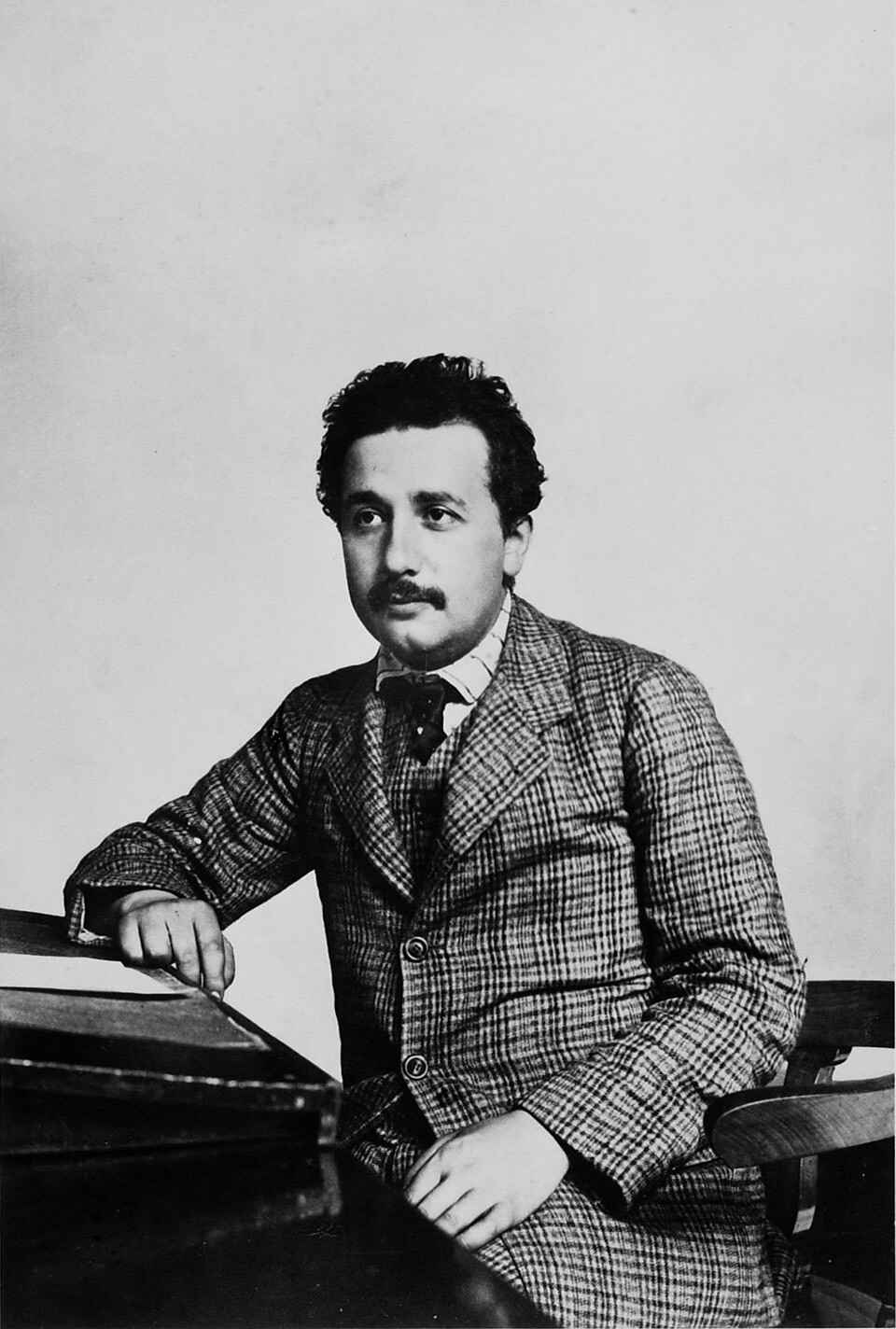 Photograph of the young Albert Einstein standing beside a desk in a suit, with dark curly hair and a mustache