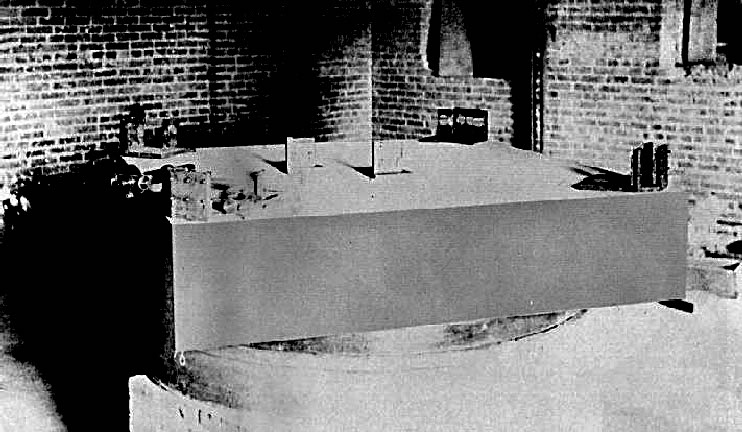 Black and white photograph of a large square stone slab carrying optical parts, resting on a round trough in a brick-walled basement