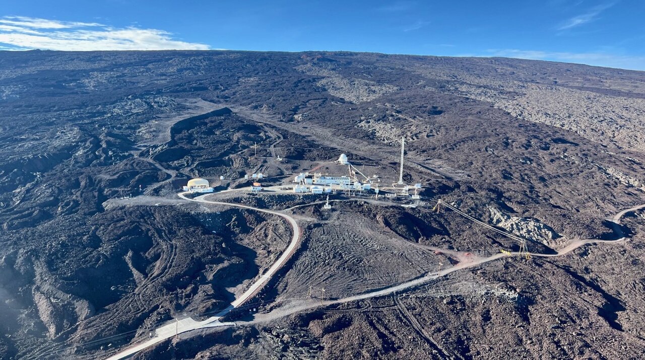 Aerial photograph of a small group of white buildings and a tall mast on a bare slope of dark lava, under a clear blue sky
