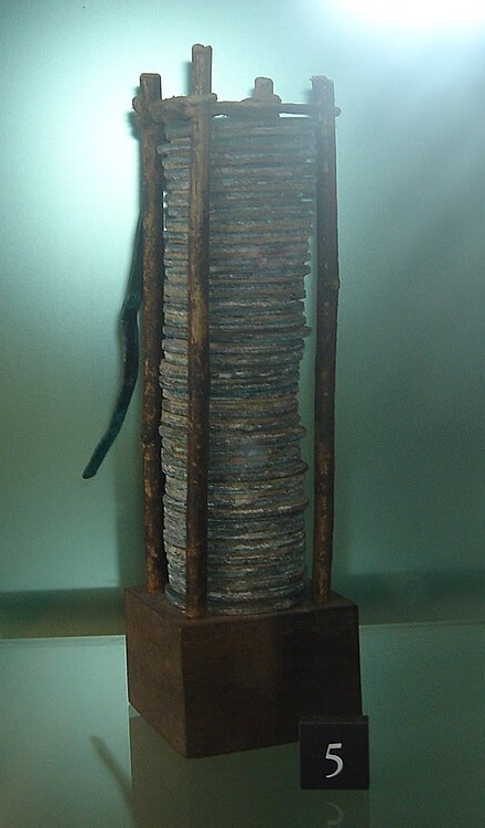 A tall stack of metal disks held upright between four thin rods on a wooden base, in a museum case