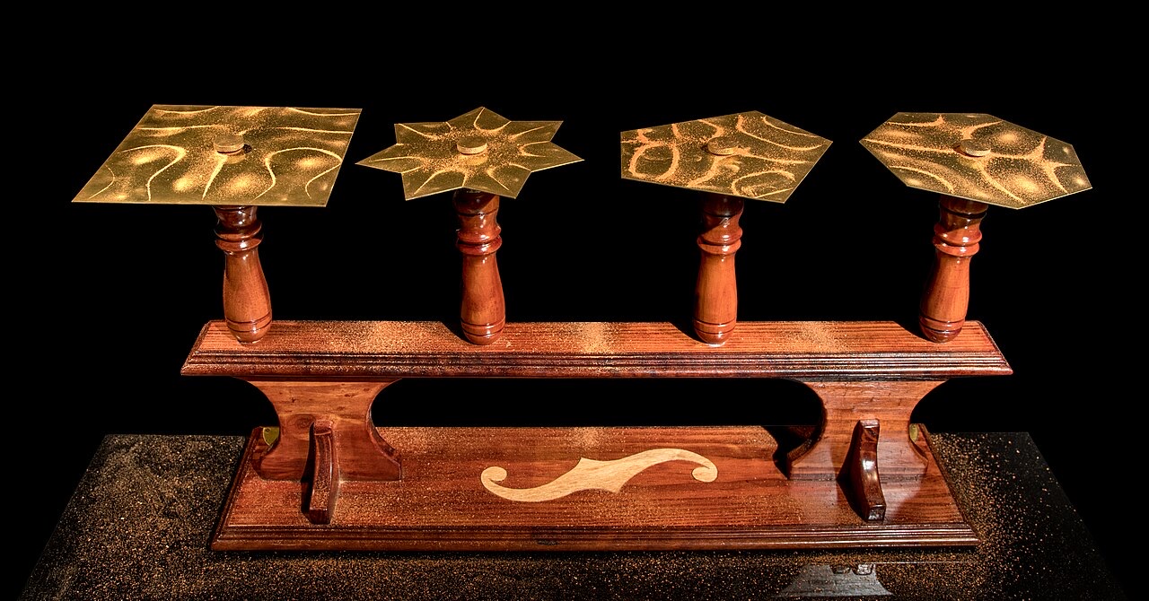 Four brass plates of different shapes on wooden stands, each covered with a pattern of curved lines of sand