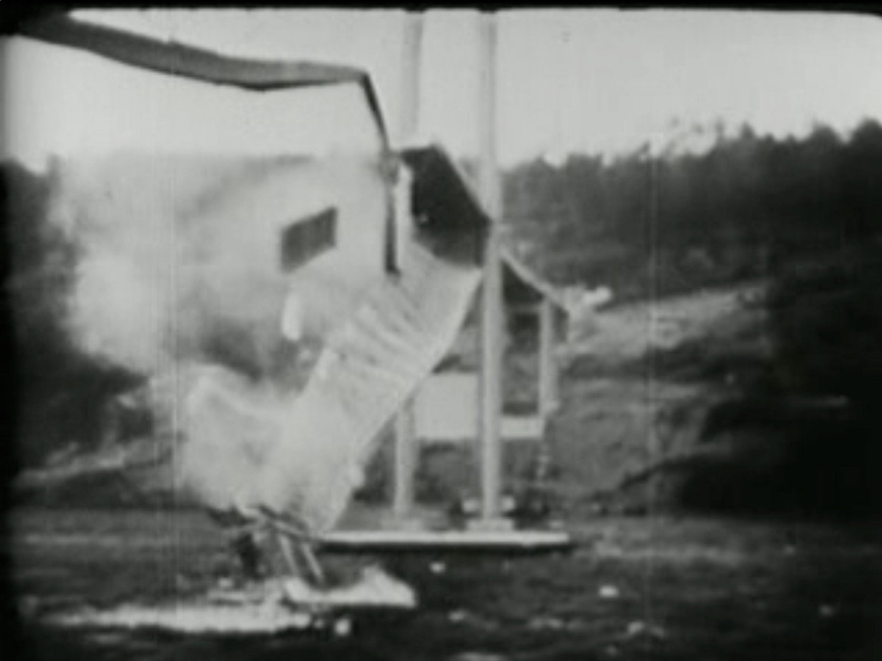 A frame of black and white film showing the roadway of a suspension bridge breaking and falling into the water between its towers