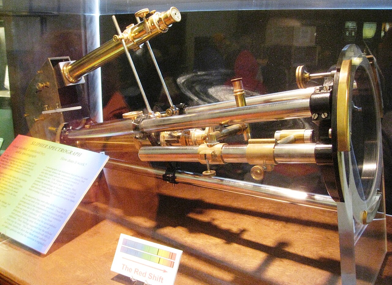 A brass spectrograph of tubes and a small telescope mounted on a frame, in a glass museum case