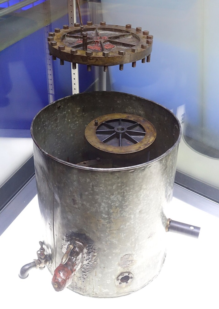 Museum display of a dull metal drum about the size of a bucket, with small windows and taps in its side, and a round brass plate with a ring of bolts held above it