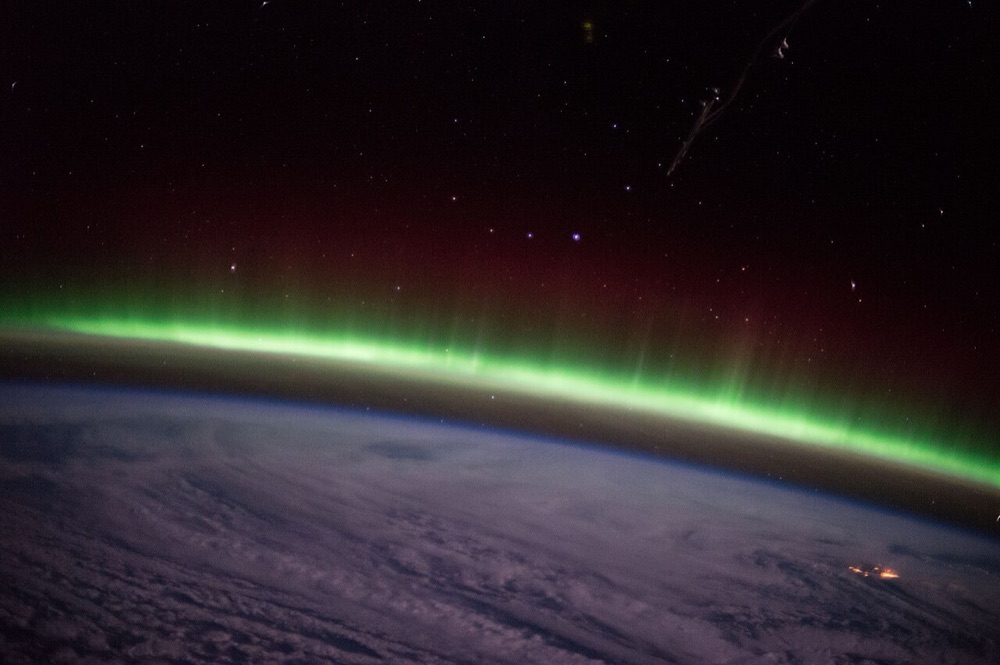 Photograph from orbit of the curved edge of the Earth at night, with a glowing green band of aurora above the clouds and part of the space station in the foreground