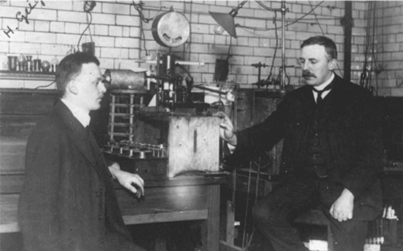 Two men in dark suits sitting either side of a bench of laboratory apparatus