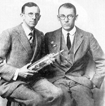 Two men in suits sitting side by side, one holding a glass vacuum tube