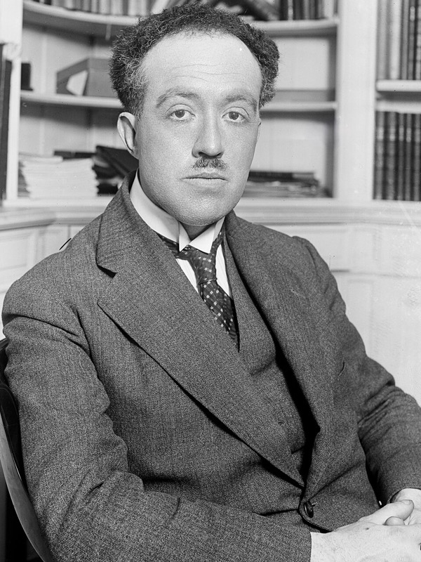 Louis de Broglie in a suit, sitting in front of bookshelves