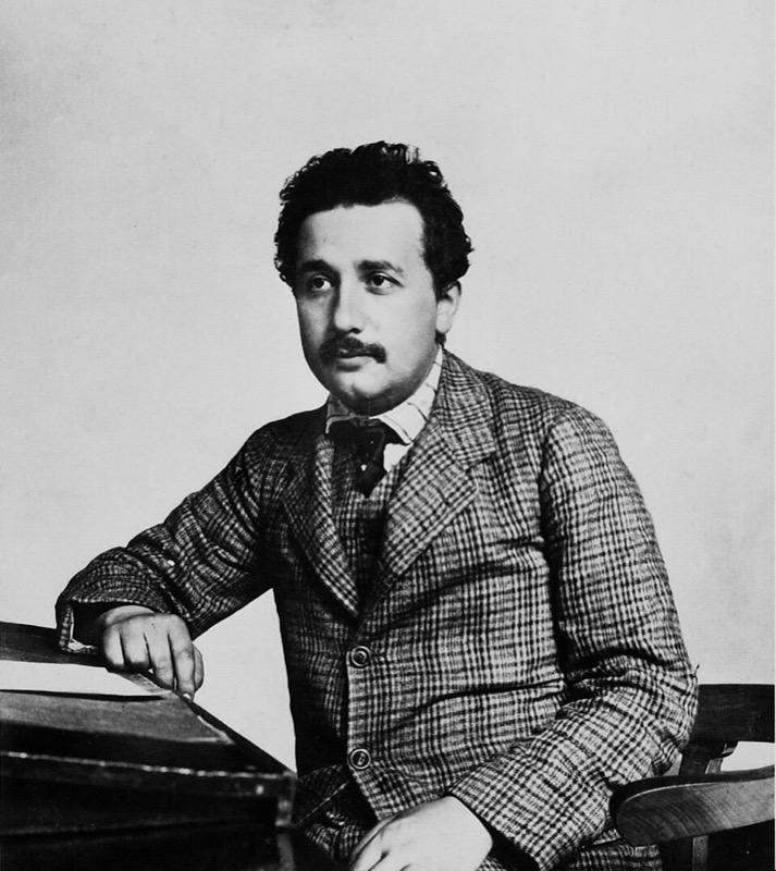 A young Albert Einstein in a checked suit, sitting beside a desk