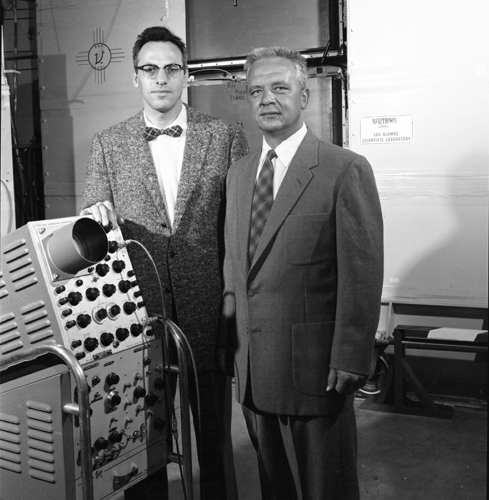 Two men in shirts and ties working at a large bank of electronic instruments covered in dials and cables