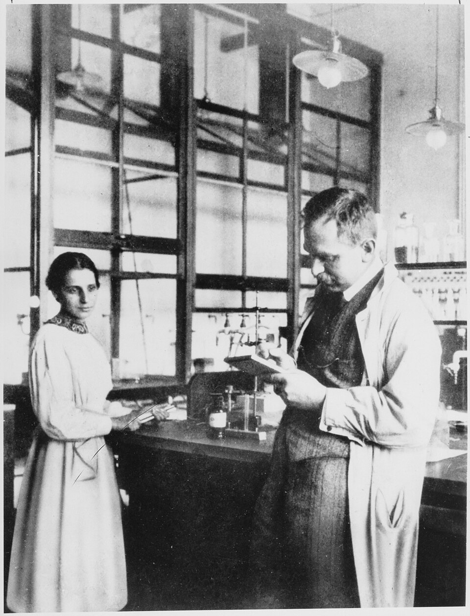 Black and white photograph of Lise Meitner and Otto Hahn standing at a laboratory bench in front of tall windows