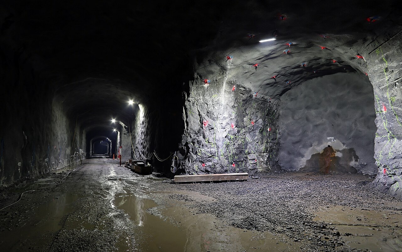 A wide tunnel cut through bare rock, lit by lamps, with a side chamber opening on the right and a wet gravel floor