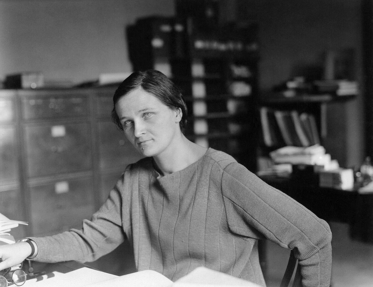 Black and white photograph of Cecilia Payne seated at a desk with papers, in front of filing cabinets and shelves